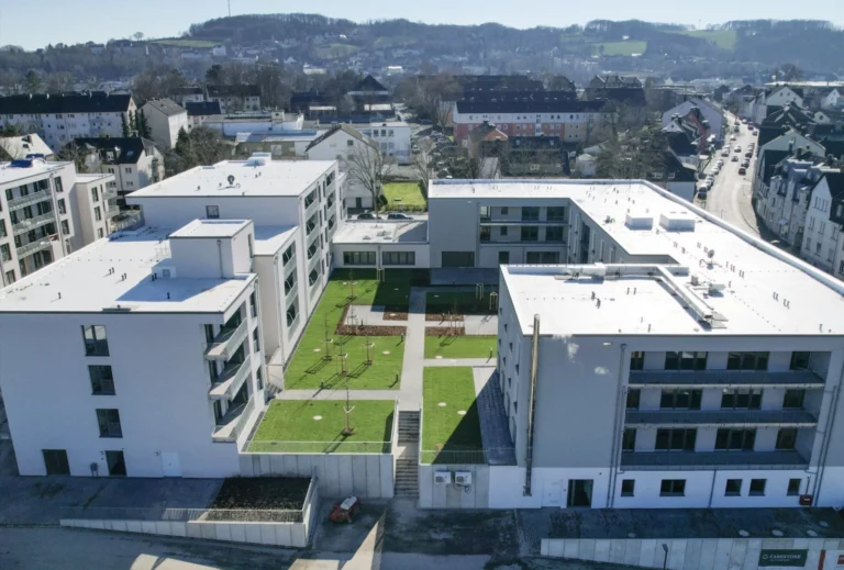 Eine Luftaufnahme eines Komplexes aus mehreren weißen, modernen Gebäuden mit flachen Dächern, die einen begrünten Innenhof umschließen. Der Innenhof hat Rasenflächen, kleine Bäume und Wege. Im Hintergrund ist eine Stadt mit Wohnhäusern und Straßen zu sehen, sowie bewaldete Hügel unter einem klaren Himmel.
