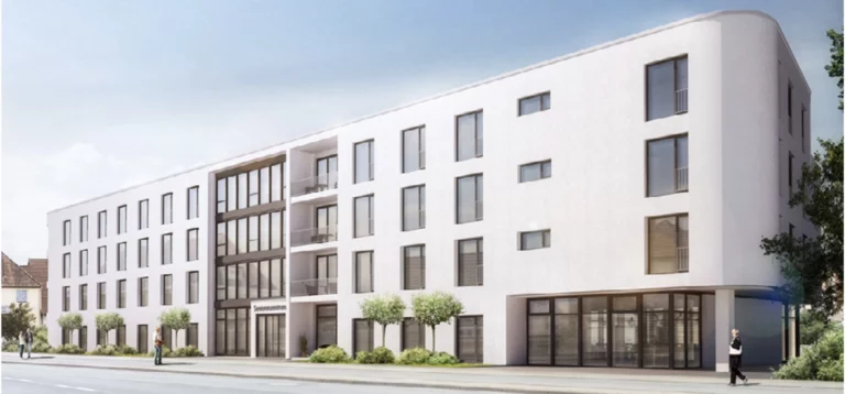 Ein moderner, weißer Wohnbau mit flacher Dachlinie und großen Balkonen, die mit Glasgeländern ausgestattet sind. Die Fassade besteht aus glatten weißen Wänden mit zahlreichen rechteckigen Fenstern. Im Erdgeschoss befindet sich eine große Glasfront, die den Eingangsbereich markiert. Ein paar kleine Bäume und Personen sind vor dem Gebäude auf einem gepflasterten Gehweg zu sehen. Der Himmel ist klar und blau.