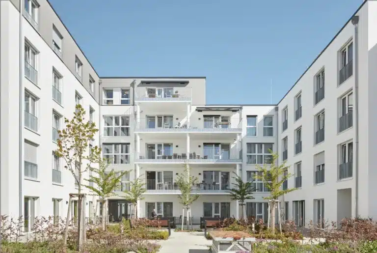 Ein moderner, weißer Wohnbau mit mehreren Stockwerken, Balkonen mit weißen Geländern und großen Fenstern. Der Innenhof ist mit Bäumen, Sträuchern und gepflasterten Wegen gestaltet, und bietet Sitzmöglichkeiten. Die Gebäude sind symmetrisch angeordnet, und der Himmel ist klar und blau.