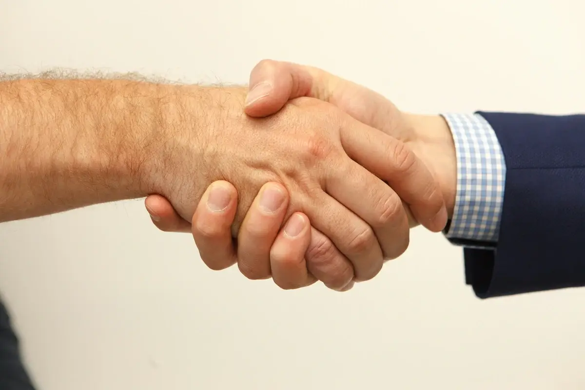 Zwei Personen geben sich die Hand, eine in einem blauen Anzug mit kariertem Hemd und die andere mit freiem Unterarm. Das Bild symbolisiert Vertrauen, Zusammenarbeit und Vertragsabschlüsse.