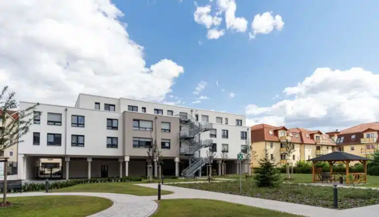 Ein moderner, weißer Wohnbau mit mehreren Stockwerken, einer Außentreppe aus Metall und rechteckigen Fenstern mit schwarzen Rahmen. Die Fassade ist hell und kombiniert mit einer grauen Akzentzone. Vor dem Gebäude erstreckt sich eine Grünanlage mit gepflegtem Rasen, Bäumen und einem gepflasterten Weg. Ein Pavillon und weitere Gebäude mit roten Dächern sind im Hintergrund sichtbar, unter einem blauen Himmel mit weißen Wolken.