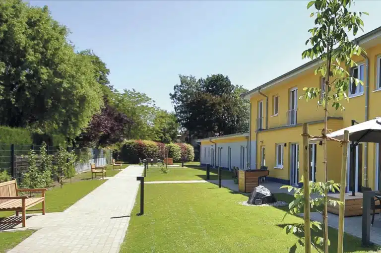 Ein gepflegter Gartenbereich vor einem modernen, gelben Wohngebäude mit mehreren Etagen, Balkonen und großen Fenstern. Der Garten hat einen gepflasterten Gehweg, der durch eine grüne Wiese mit Bäumen und Büschen führt. Auf dem Weg stehen hölzerne Bänke, und am rechten Rand befindet sich ein junger Baum, der mit Stützen fixiert ist. Im Hintergrund sind weitere Bäume und Sträucher zu sehen, und der Himmel ist klar und blau. Ein Sonnenschirm und Gartenmöbel sind auf einer Terrasse des Gebäudes sichtbar.