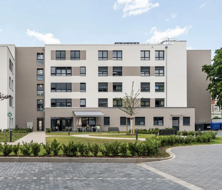 Ein moderner, mehrstöckiger Wohnbau mit einer Fassade aus weißem Putz und grauen Akzenten. Das Gebäude hat zahlreiche Fenster und einen Sitzbereich mit Tischen und Stühlen im Erdgeschoss, der von einem gepflegten Garten mit Büschen und einem Baum umgeben ist. Ein gepflasterter Weg führt zum Gebäude, und der Himmel ist klar und blau.