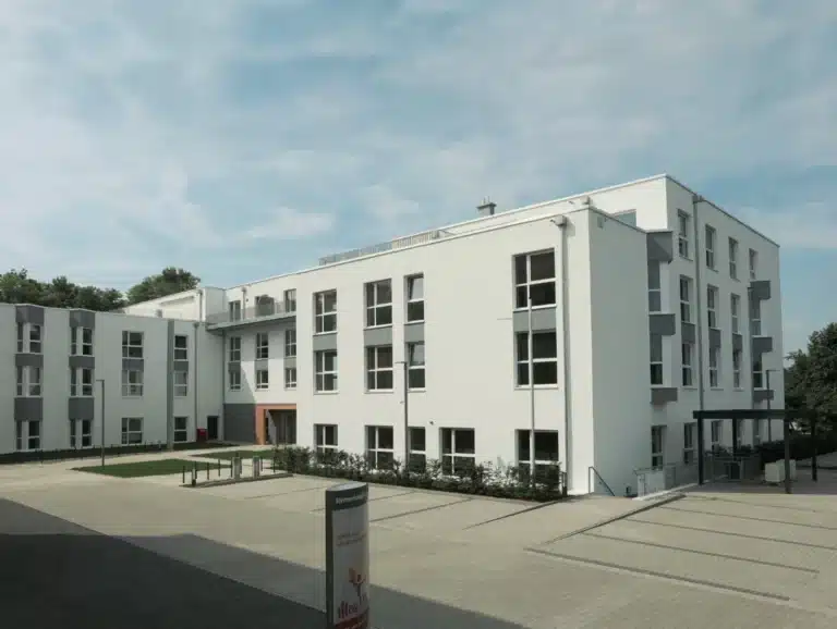 Ein modernes, mehrstöckiges Wohngebäude mit weißer Fassade und grauen Akzenten, das einen gepflasterten Vorplatz mit einem Eingang umgibt. Das Gebäude hat mehrere Etagen mit großen Fenstern und Balkonen, umgeben von Bäumen und gepflegten Büschen. Ein Schild mit der Aufschrift „Seniorenresidenz“ steht vor dem Gebäude, und der Himmel ist teilweise bewölkt.