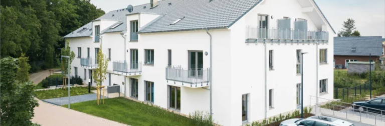 Ein moderner, weißer Wohnbau mit mehreren Stockwerken, Balkonen mit Metallgeländern und großen Fenstern. Die Fassade ist glatt und hell, und das Gebäude ist von einem gepflegten Garten mit grünen Bäumen, Sträuchern und einem gepflasterten Weg umgeben. Ein Auto parkt auf dem Platz vor dem Gebäude, und der Himmel ist klar mit einigen Wolken.
