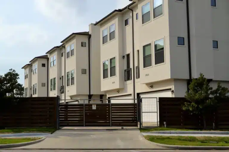 Eine Reihe moderner, mehrstöckiger Gebäude mit beige-weißer Fassade und dunklen Dächern, die entlang einer Straße stehen. Die Gebäude haben Garagen mit Toren und Balkone, umgeben von einem hölzernen Zaun und einigen Bäumen. Der Himmel ist teilweise bewölkt, und ein gepflegter Rasenstreifen grenzt an die Straße.