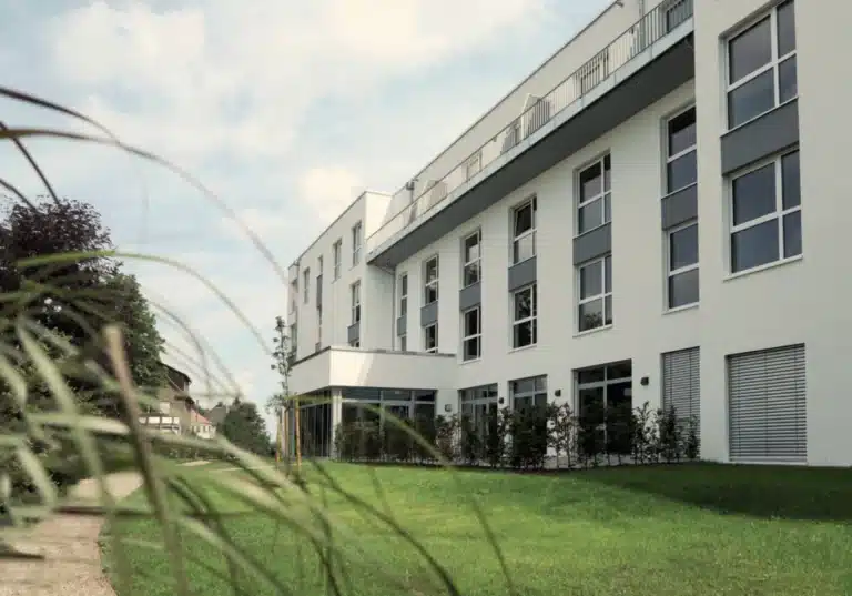 Ein modernes, mehrstöckiges Wohngebäude mit weißer Fassade und grauen Akzenten, das von einer grünen Wiese und Bäumen umgeben ist. Das Gebäude hat mehrere Etagen mit großen Fenstern und Balkonen. Im Vordergrund ist eine gepflegte Grünanlage mit Büschen und einem Baum zu sehen, und der Himmel ist teilweise bewölkt.