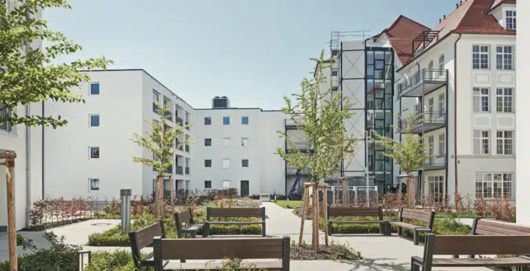 Ein Komplex aus modernen, weißen Gebäuden mit mehreren Etagen, die einen gepflasterten Innenhof umschließen. Der Innenhof hat Bänke, kleine Bäume und gepflegte Grünanlagen. Im Hintergrund sind weitere Gebäude mit traditioneller Architektur zu sehen, und der Himmel ist klar und blau.